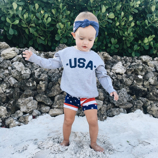 A young child stands barefoot on sand by rocks, wearing the USA Pullover • Kids! sweatshirt, star-spangled shorts, and a blue headband with a bow. Green bushes fill the background.
