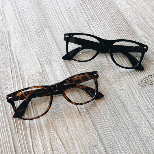 Two pairs of eyeglasses on a light wooden surface: one pair is Nerd Glasses (kids) with classic black frames, while the other features a brown tortoiseshell pattern.