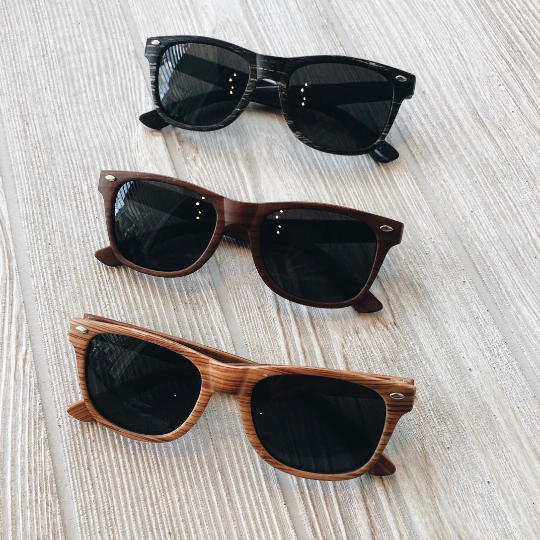 Three pairs of Marbled Sunnies • Kids sunglasses with dark lenses and black, dark brown, and light brown wood-textured frames, arranged vertically on a light wooden surface—your must-have final sale accessory.