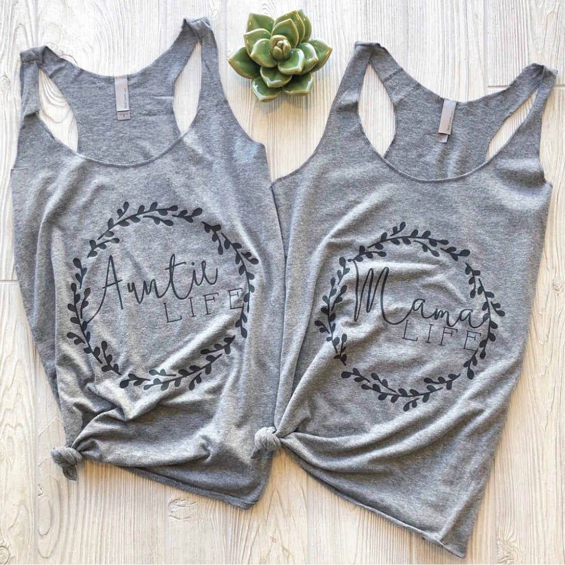 Two gray womens tank tops, including the Auntie Wreath Tank with an Auntie Life wreath design and another reading Mama Life, both with leafy details, are laid flat on a light wood surface beside a small succulent.