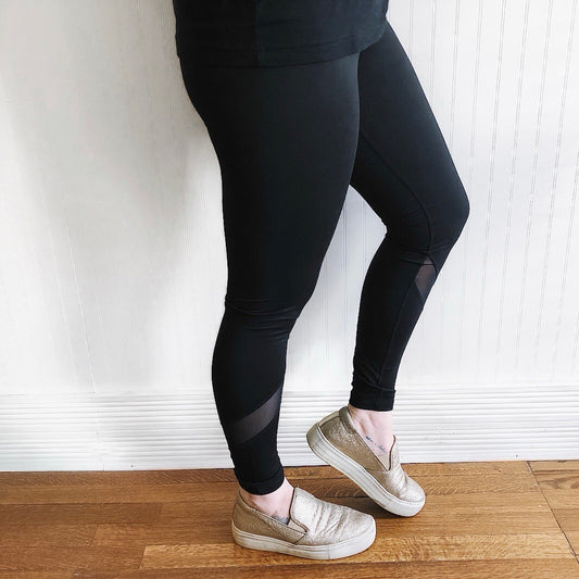 Model wears Mesh Splice Leggings • 1324—black leggings with athletic detailing and mesh panels—paired with metallic slip-on shoes, standing on a wooden floor against a white wall. Only the lower body is shown.