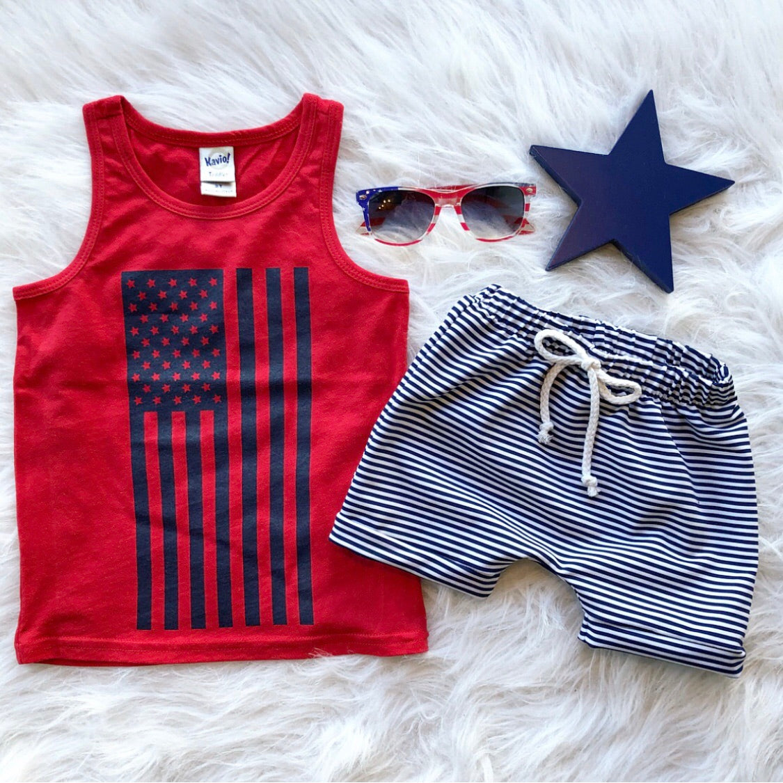 A flat lay of the Flag Tank • Kids—a super-soft red tank top with a vertical blue American flag—paired with blue and white striped drawstring shorts, star-shaped sunglasses, and a navy star on white faux fur.