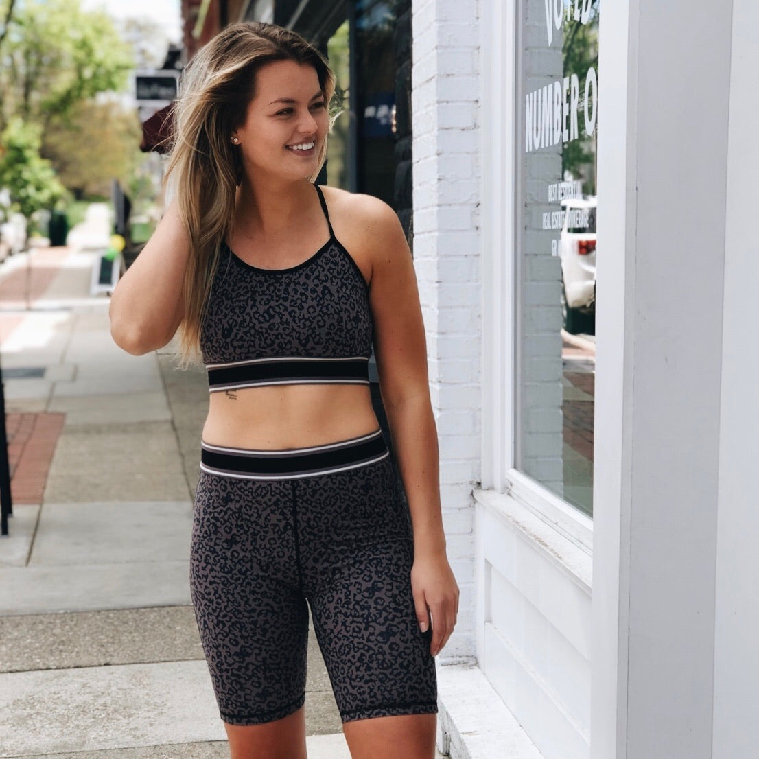 A woman smiles on a sunny sidewalk, looking relaxed in matching black and grey leopard print biker shorts and the Cheetah Sports Bra—true to size and ultra comfy—with one hand in her hair.