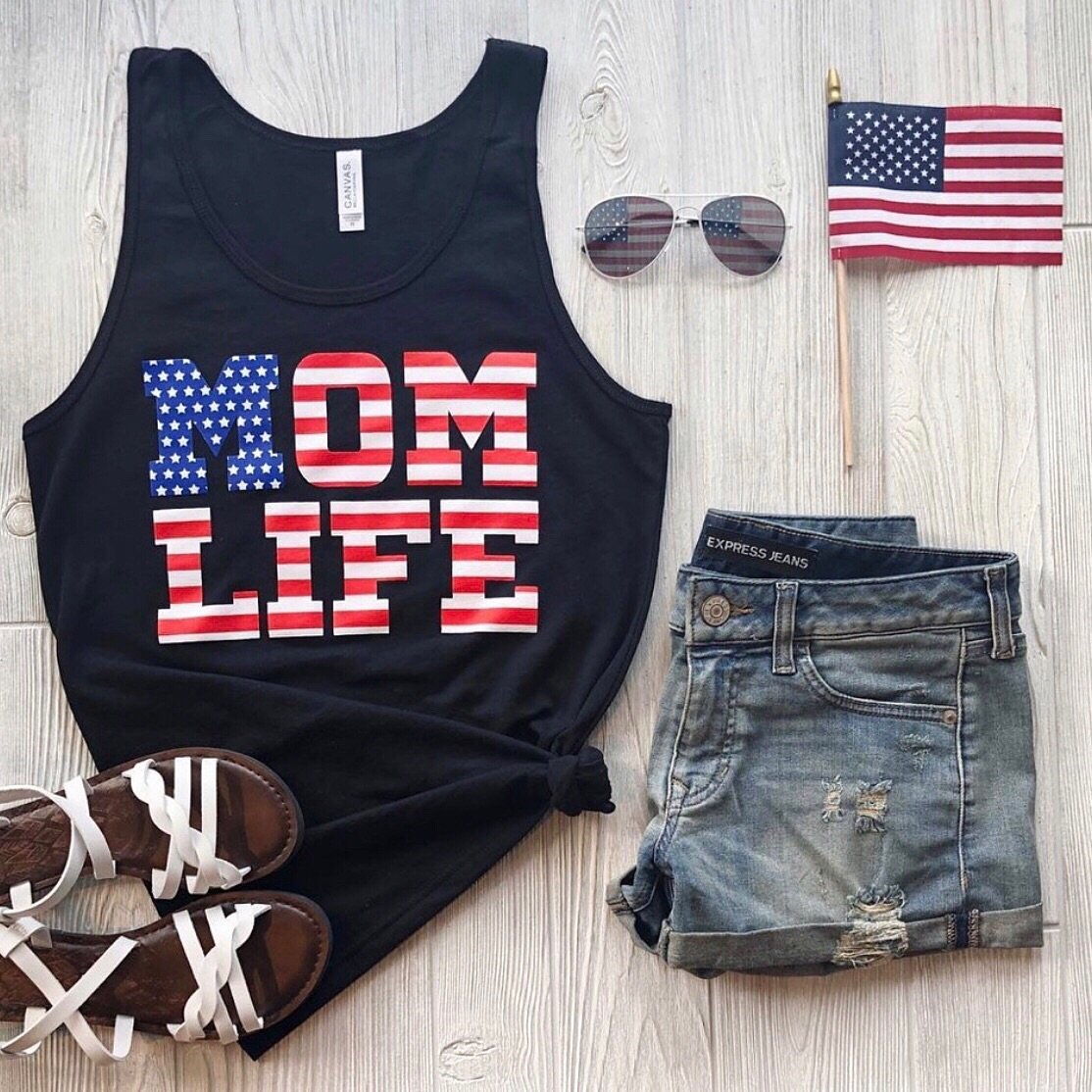 Flat lay of summer outfit: Mom Flag Print • Unisex Tank Top, denim shorts, brown sandals, American flag sunglasses, and a small American flag on a light wooden background.