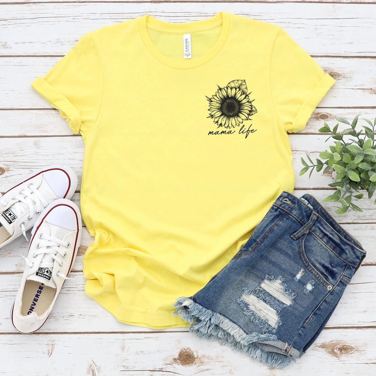 The Mama Life • Sunflower Tee is a yellow Bella Canvas shirt with a black sunflower and mama life text, styled with denim shorts, white sneakers, and a small green plant on a white wooden surface.