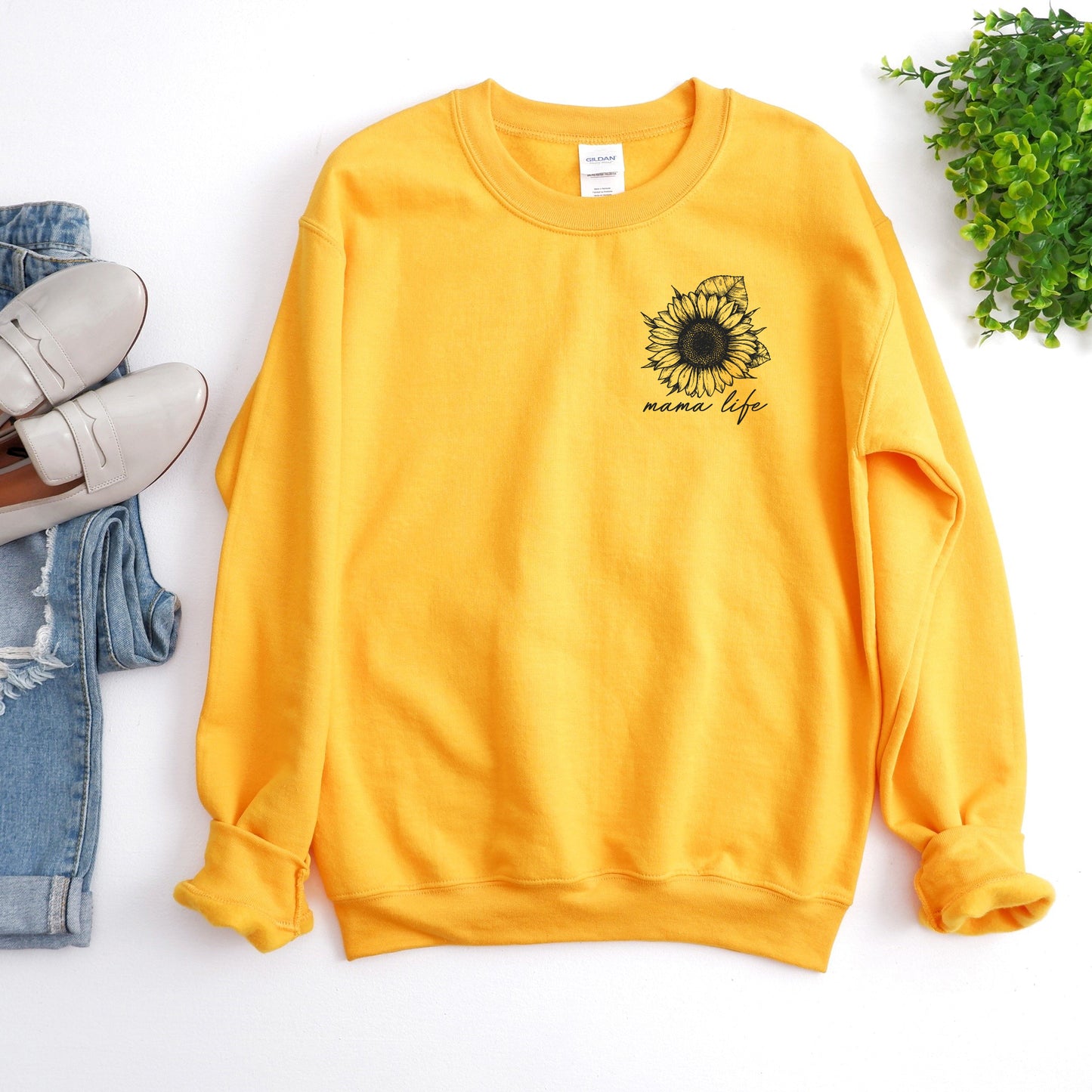The Sunflower • Gold Pullover is a yellow crewneck sweatshirt with a black sunflower mama life graphic, unisex fit. Displayed flat next to light blue jeans, beige loafers, and a small green plant.