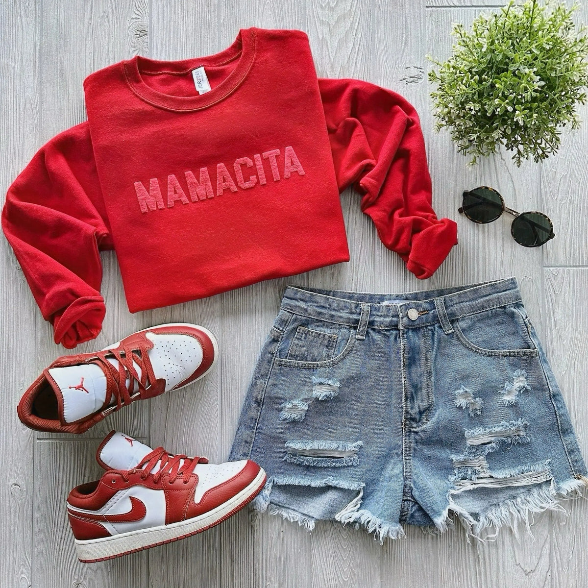 A red sweatshirt with MAMACITA text, distressed denim shorts, red and white sneakers, round black sunglasses, and a small green plant arranged on a light wood surface.