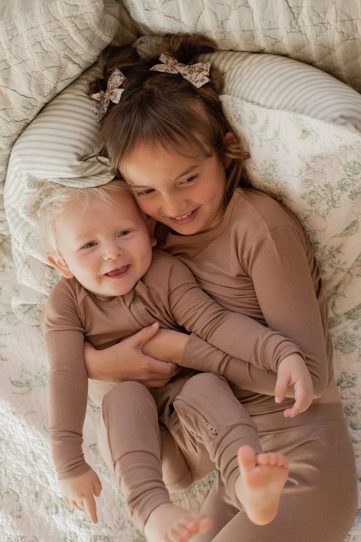 Two young children in matching beige pajamas cuddle and smile joyfully while lying on a cozy, patterned bed with light-colored bedding.