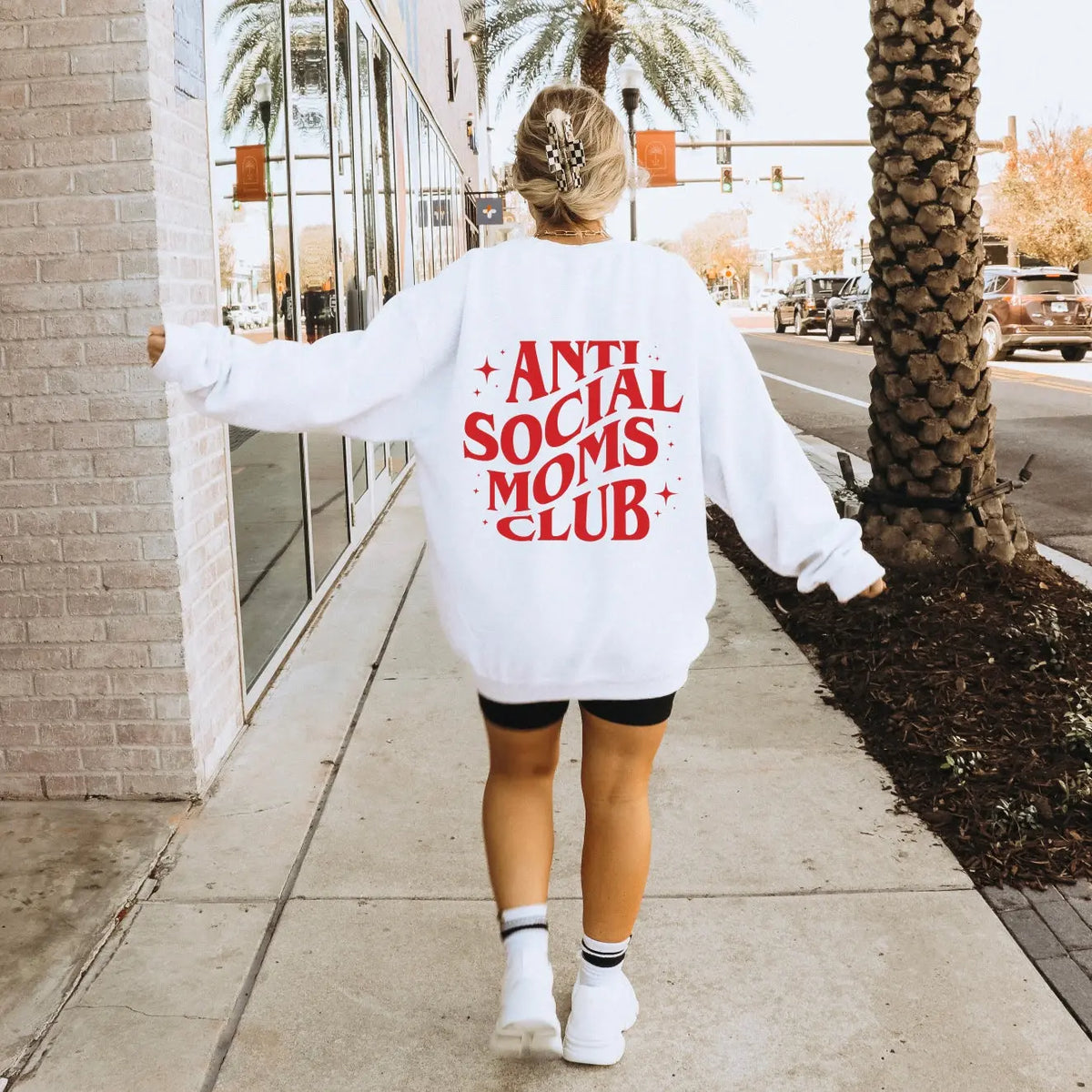 A woman walks down a sunny sidewalk wearing a white sweatshirt that says ANTI SOCIAL MOMS CLUB in bold red letters on the back.