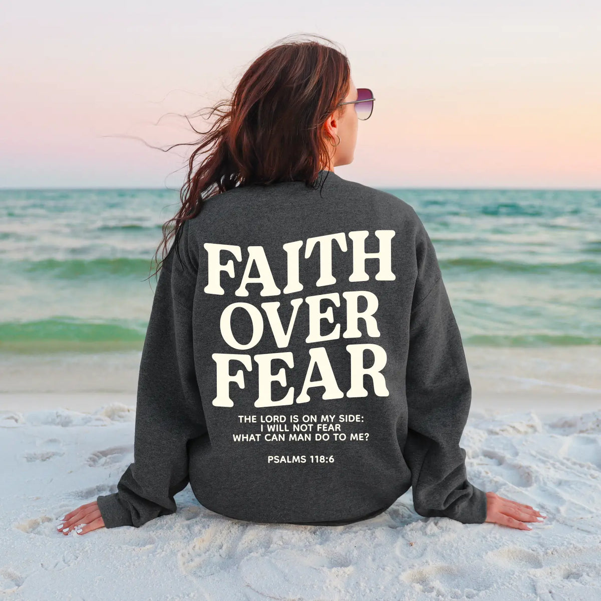 A woman with long hair sits on the sand at the beach, wearing sunglasses and a sweatshirt that reads, FAITH OVER FEAR. The Lord is on my side; I will not fear. Psalms 118:6.