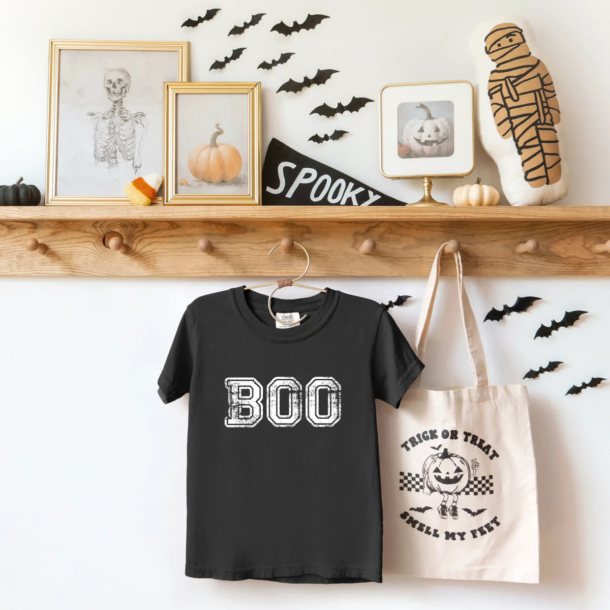 A Halloween-themed display with framed art, bat cutouts, a pumpkin, a mummy pillow, a BOO t-shirt, and a tote bag reading Trick or Treat, Smell My Feet hanging on a wooden rack.