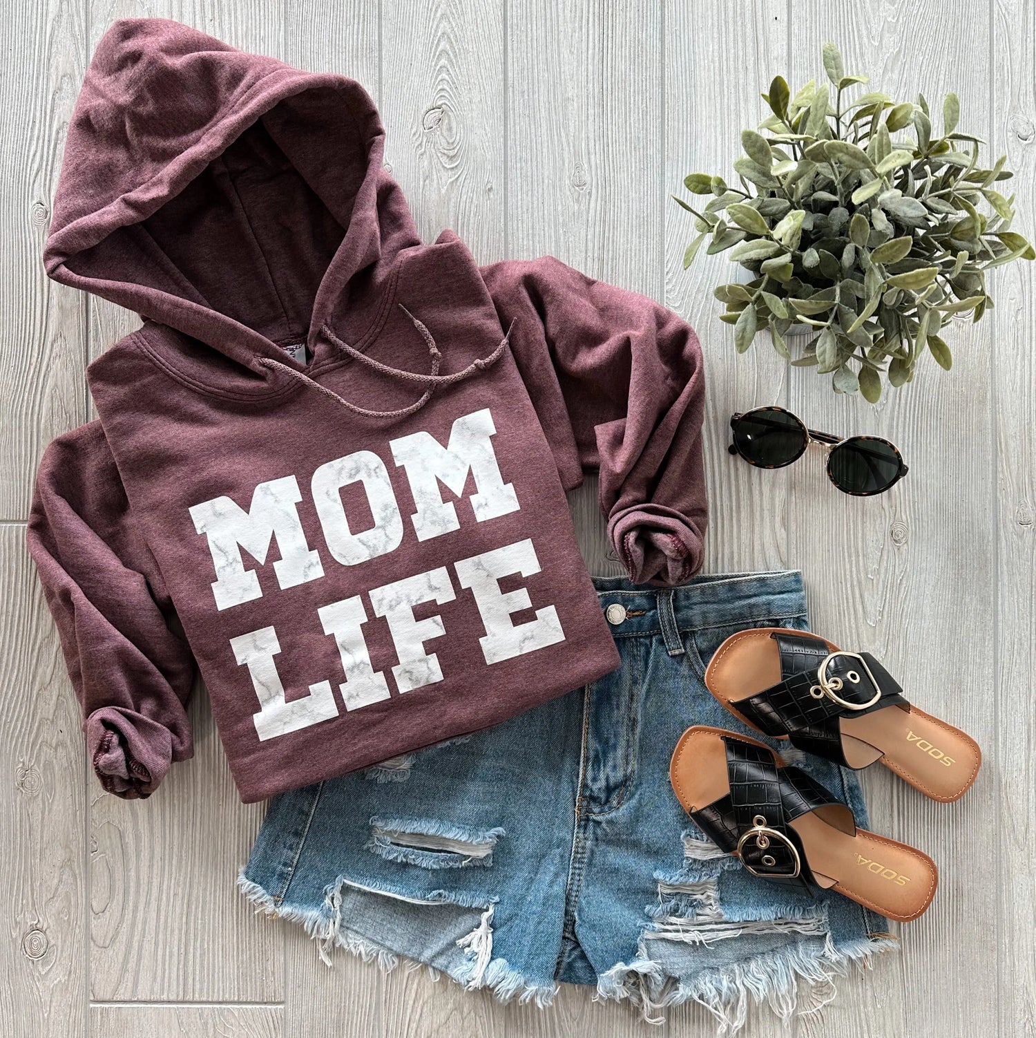 A flat lay of a maroon hoodie with MOM LIFE text, denim shorts, black sandals, black sunglasses, and a small potted plant on a light wood background.