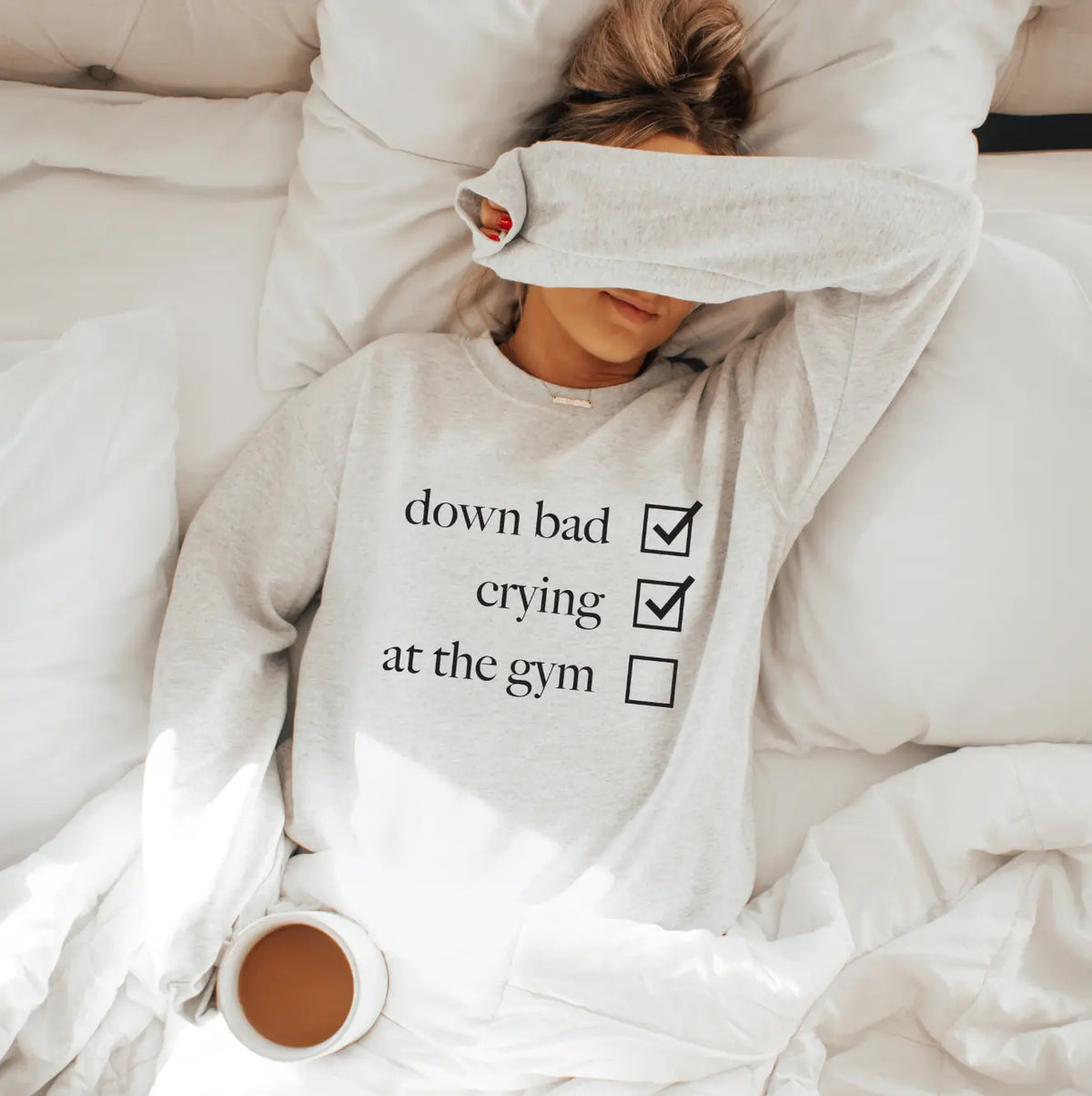 A person lies in bed with their arm over their face, wearing the Not at the Gym • Ash Pullover—a cotton/polyester unisex sweatshirt featuring check boxes for down bad and crying. A mug of coffee rests on the white blanket beside them.