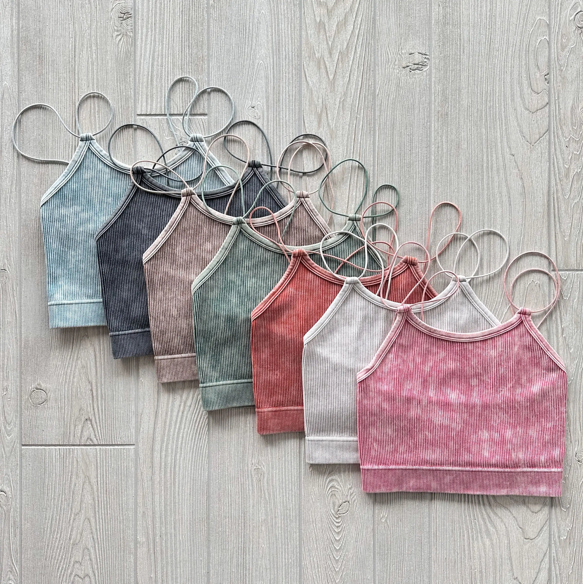 Seven Caiti Vintage Crop halter tops in blue, gray, brown, taupe, green, light pink, and bright pink are displayed side by side on a light wood floor, highlighting their ribbed design and soft, stretchy fabric.