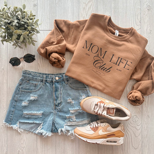 A flat lay of a casual outfit featuring a caramel "Mom Life Club • Caramel Pullover," distressed denim shorts, beige and pink Nike sneakers, black sunglasses, and a small green plant on a light wood surface.