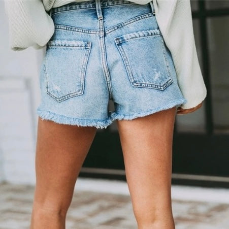 Someone wearing Myra Denim Shorts in medium wash with high waist, distressed details, and frayed hems pairs them with a light top, standing with their back to the camera.