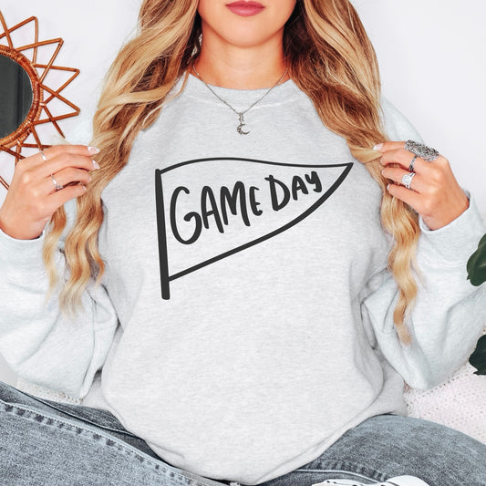 A woman with long wavy hair casually models the Game Day • Ash Pullover, a comfy unisex sweatshirt in ash gray with GAME DAY inside a pennant. She wears rings and a crescent moon necklace while holding her hair.