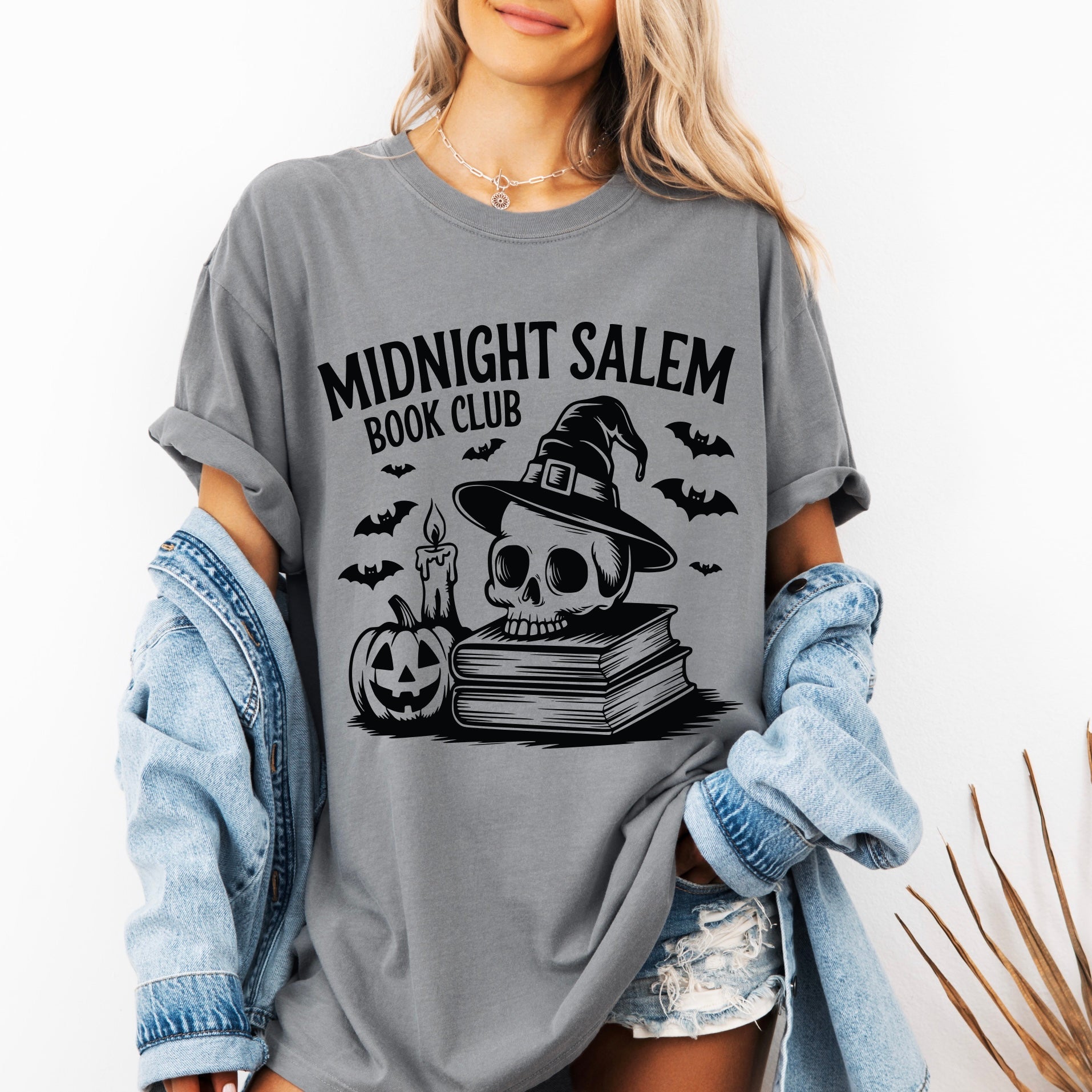 A woman wears the Midnight Salem Book Club • Comfort Tee in gray, showcasing a skull with witch hat, candle, bats, stacked books, and a jack-o-lantern. She pairs this garment-dyed shirt with a denim jacket and shorts for her zombie-inspired look.