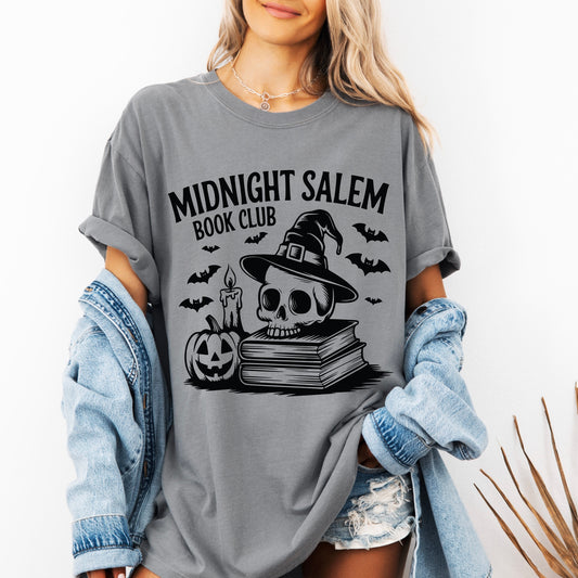 A woman wears the Midnight Salem Book Club • Comfort Tee in gray, showcasing a skull with witch hat, candle, bats, stacked books, and a jack-o-lantern. She pairs this garment-dyed shirt with a denim jacket and shorts for her zombie-inspired look.
