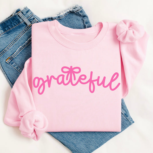 The grateful • Pink Pullover is a comfy unisex cotton-poly sweatshirt featuring "grateful" in pink cursive. Styled with pink scrunchies on the sleeves, it rests atop blue jeans for a cozy, fashionable look.