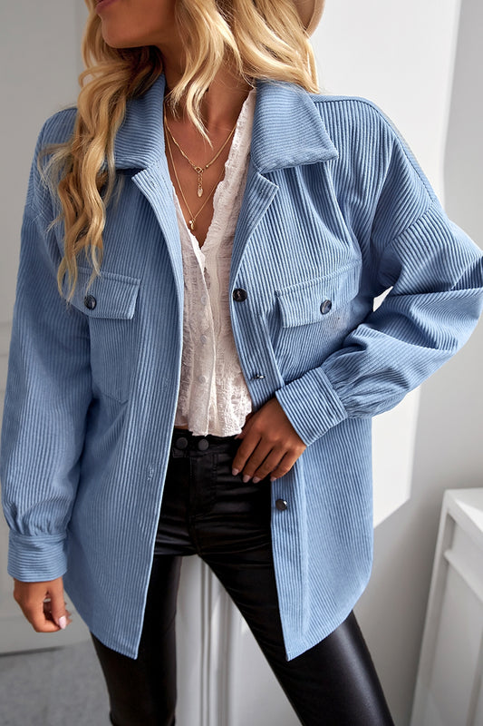 A woman with wavy blonde hair wears the oversized light blue Cassandra Shacket over a white lace top and black leather pants, accessorized with layered necklaces and a hat, standing indoors by a white wall.