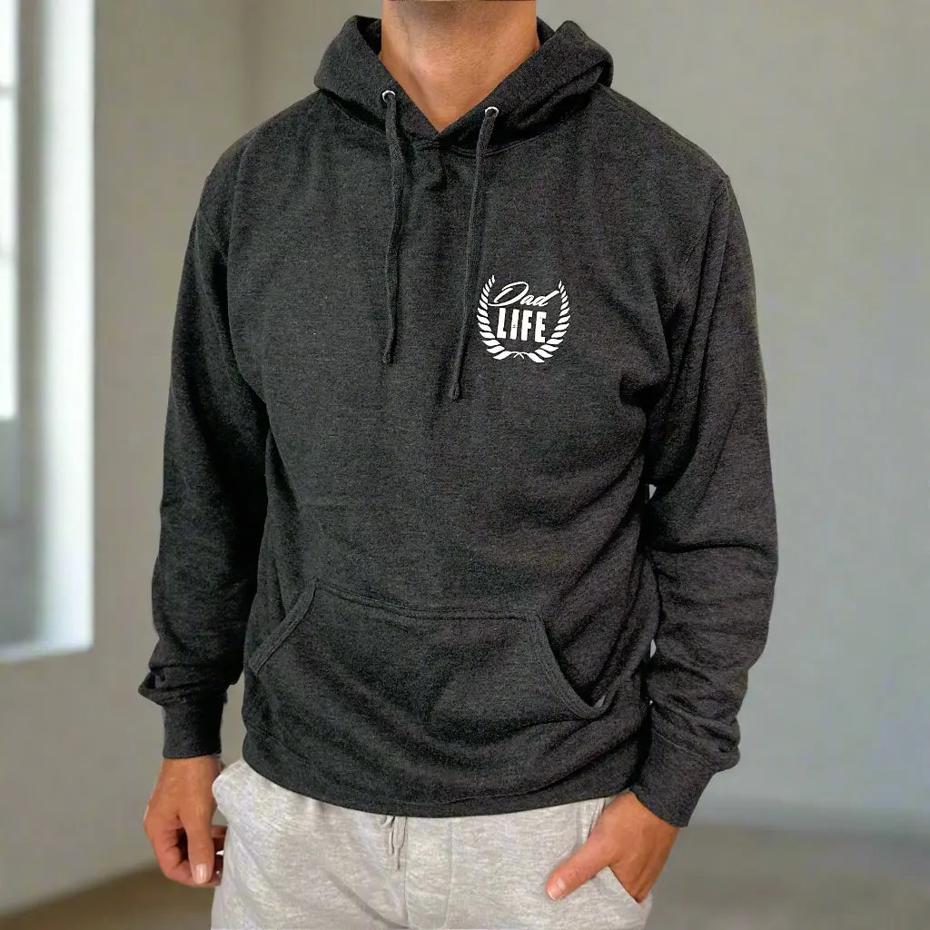 A person wears the Dad Life Crest • Heather Graphite Hoodie, featuring a white laurel on the chest, styled with light gray sweatpants indoors. This hoodie offers casual comfort and fits true to size.