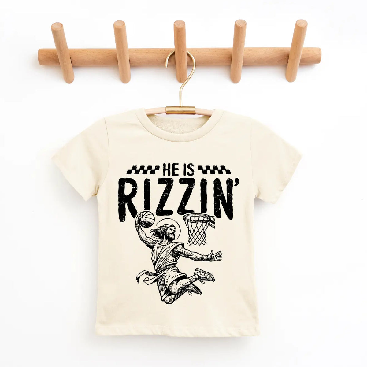 The He is Rizzin • Kids Natural Tee, in beige and hung on a wooden hanger, shows a basketball player dunking with “He is Rizzin’” above—crafted from super-soft fabric for Easter or everyday wear.
