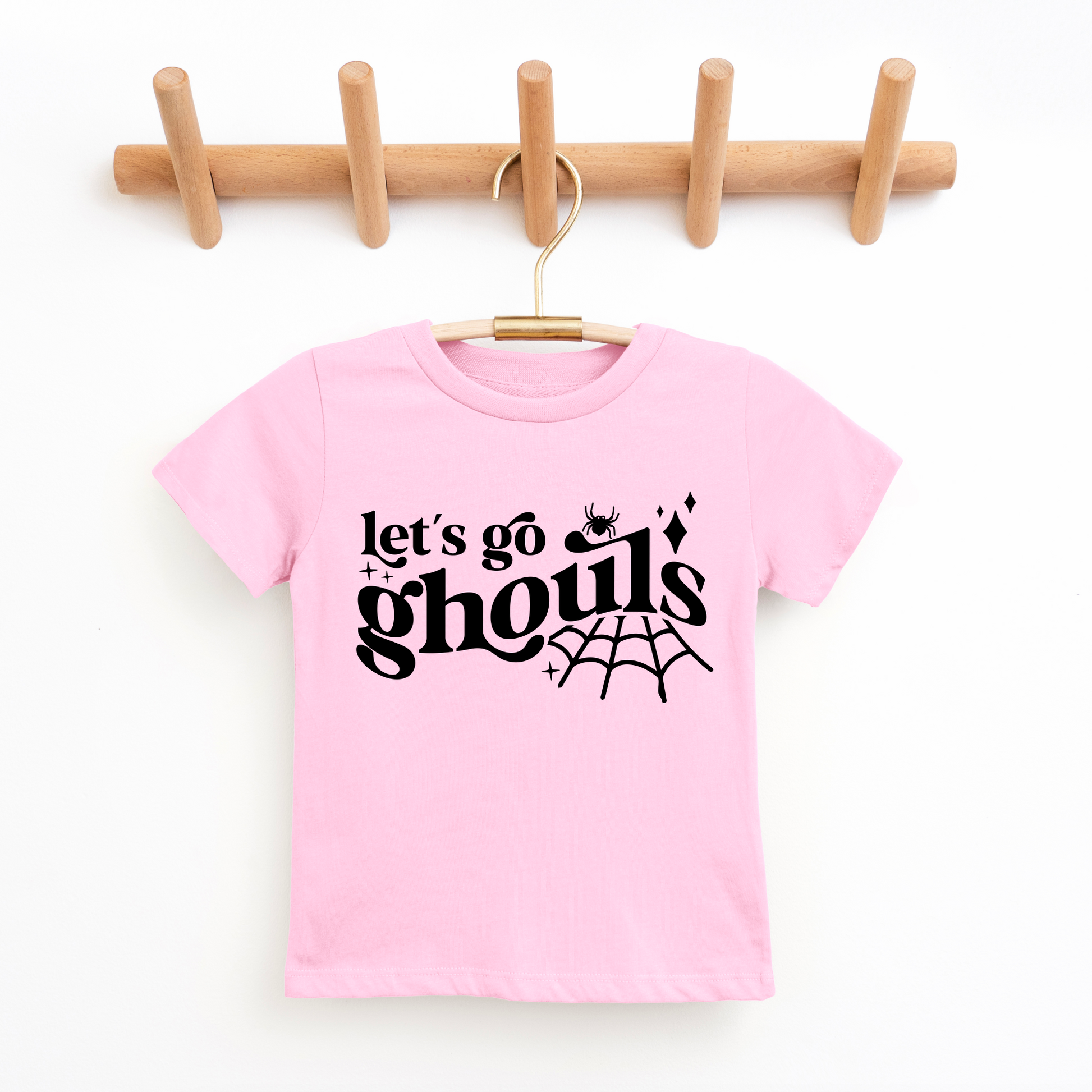 A pink Lets Go Ghouls • Kids Tee made from super-soft cotton hangs on a wooden hook rack, with black text, a small spider, sparkles, and a spiderweb—perfect for kids who love fun and spooky shirts.