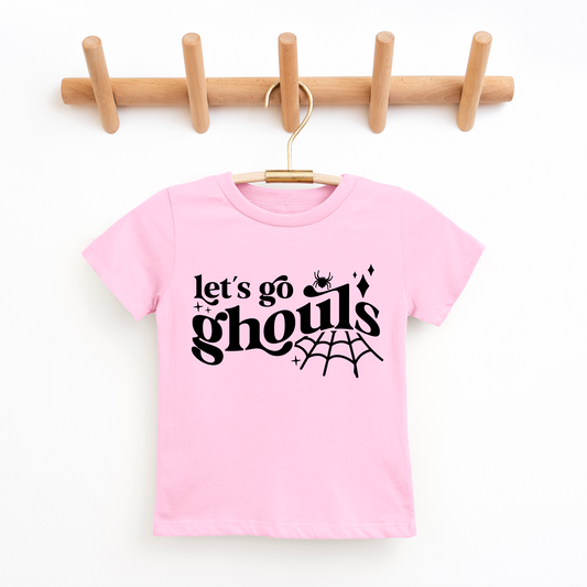 A pink Lets Go Ghouls • Kids Tee made from super-soft cotton hangs on a wooden hook rack, with black text, a small spider, sparkles, and a spiderweb—perfect for kids who love fun and spooky shirts.