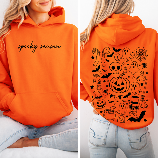 A woman wears the Spooky Season • Neon Orange Hoodie with “spooky season” in black script on the front and black Halloween-themed graphics—ghosts, pumpkins, bats, candy, and stars—on the back.