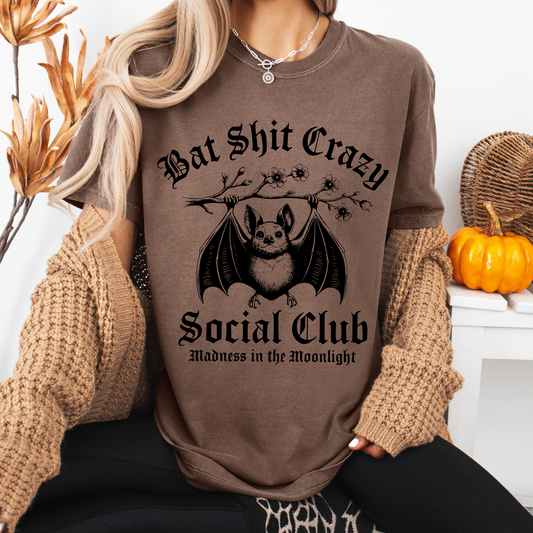 A woman wears the Crazy Social Club • Comfort Tee, a brown, garment-dyed shirt featuring a bat graphic and Bat Shit Crazy Social Club: Madness in the Moonlight—a cozy pick for fall or any zombie wardrobe.