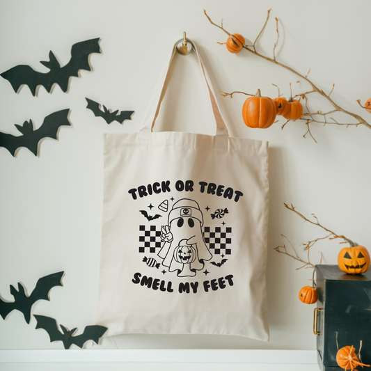 The Trick or Treat Tote, made from sustainable 100% cotton canvas, hangs on a wall with a cartoon ghost, candy, and Trick or Treat Smell My Feet text. Black paper bats and pumpkin decorations complete the Halloween look.