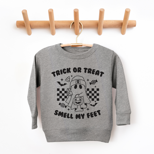 The Smell my Feet • Kids Pullover, a cozy gray unisex sweatshirt, hangs on a wooden rack. It features a cartoon ghost with candy, bats, stars, checkerboard patterns, and Trick or Treat Smell My Feet, perfect for Halloween-loving kids.