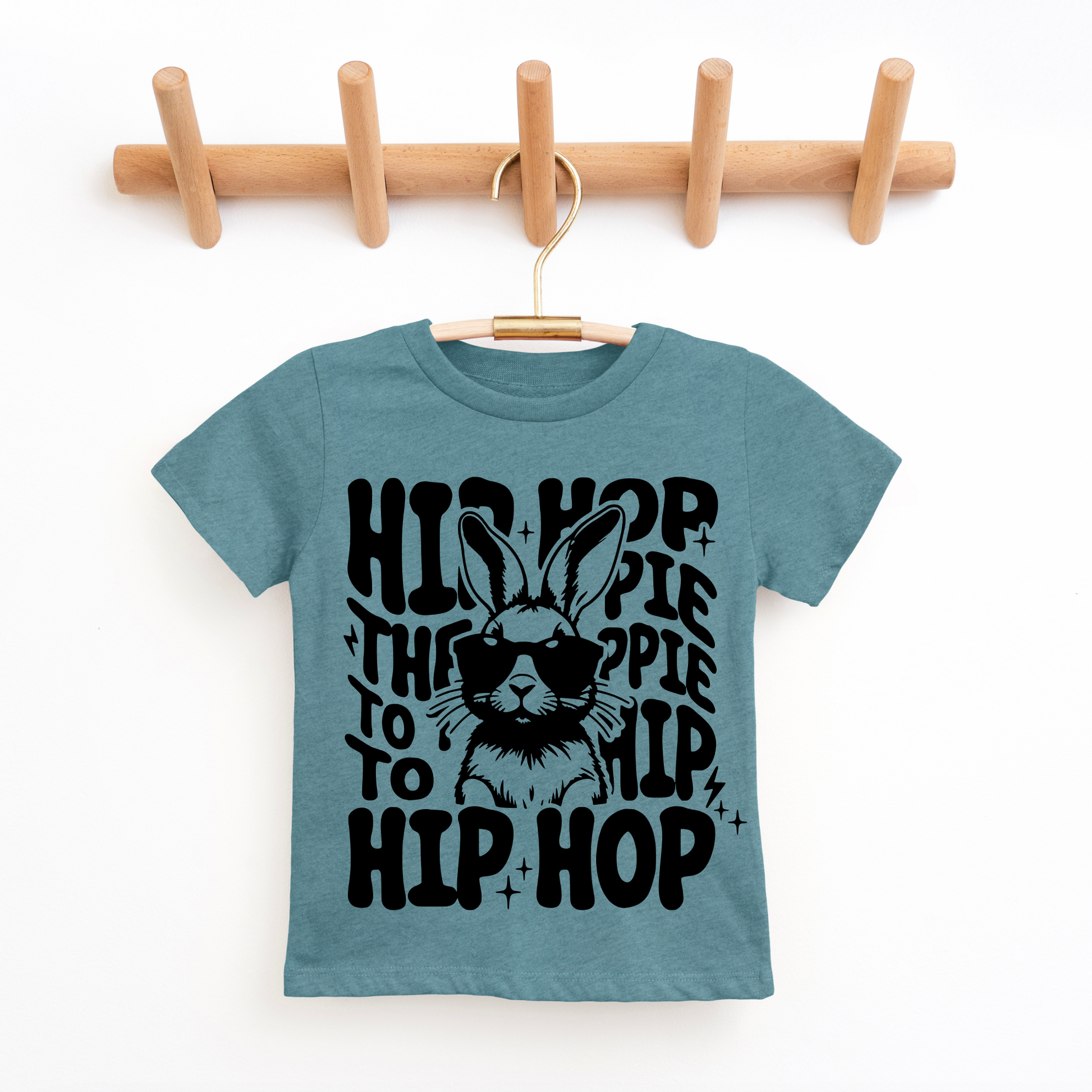 Blue t-shirt with black graphic design of a rabbit wearing sunglasses and text, hanging on a wooden rack.