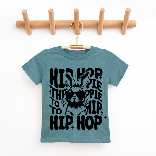 Blue t-shirt with black graphic design of a rabbit wearing sunglasses and text, hanging on a wooden rack.