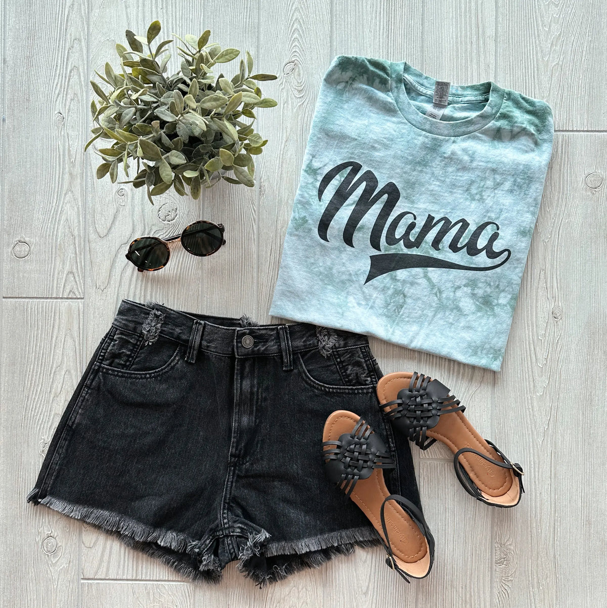 Flat lay of a casual outfit: Mama Sporty • Moss Tie Dye unisex T-shirt, black denim shorts, black fringe sandals, round sunglasses, and a small potted plant on a light wood floor.