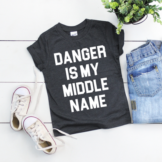The DANGER • Kids Tee, a black charcoal shirt with bold white DANGER IS MY MIDDLE NAME print, is styled alongside white sneakers, blue jeans, and a potted plant. Kavio Size Specs guarantee the perfect fit.