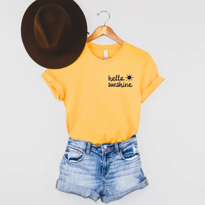 The Hello Sunshine • Gold Tee, featuring hello sunshine and a sun graphic, hangs on a wooden hanger next to denim shorts and a brown wide-brim hat against a white background.