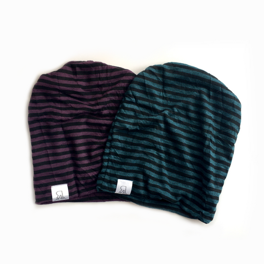 Two Striped Beanies, one in dark purple/black and the other in dark green/black, feature horizontal stripes and a small white logo tag at the edge. Made of comfortable interlock knit for a stylish look, shown on a white background.