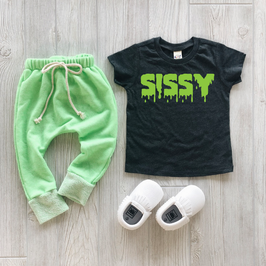 A toddler outfit on a wooden floor features light green jogger pants with a drawstring, white slip-on shoes with black labels, and a Kavio brand Sissy • Slime Print Tee in black with SISSY in bright green.