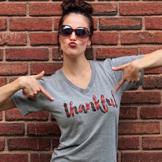 A woman in sunglasses and a gray Thankful Plaid • Tee poses playfully before a brick wall, pointing at the shirt’s design. This comfy unisex tee is crafted from a soft poly/cotton blend and features a fun “Thankful” plaid graphic.