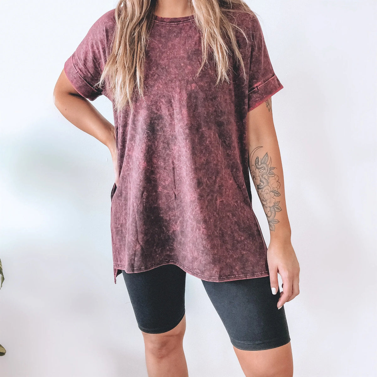 A person stands against a plain background, wearing a faded maroon oversized T-shirt and high waisted Brushed Biker Shorts with an elastic waistband. A floral tattoo is visible on their left forearm, and their face is not shown.