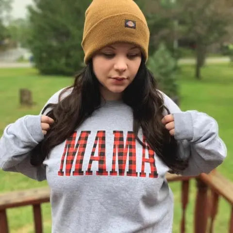 A woman stands outside on a wooden deck, surrounded by green trees and grass, wearing a tan beanie and the MAMA • Plaid Print Pullover—a gray unisex sweatshirt featuring MAMA in red plaid letters.