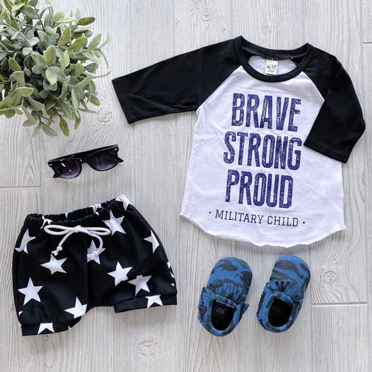 A flat lay showcases the Military Kid Raglan—a white and black shirt with “Brave Strong Proud Military Kid” in blue—paired with black star shorts, blue camo shoes, sunglasses, and a green plant on a light wooden floor.