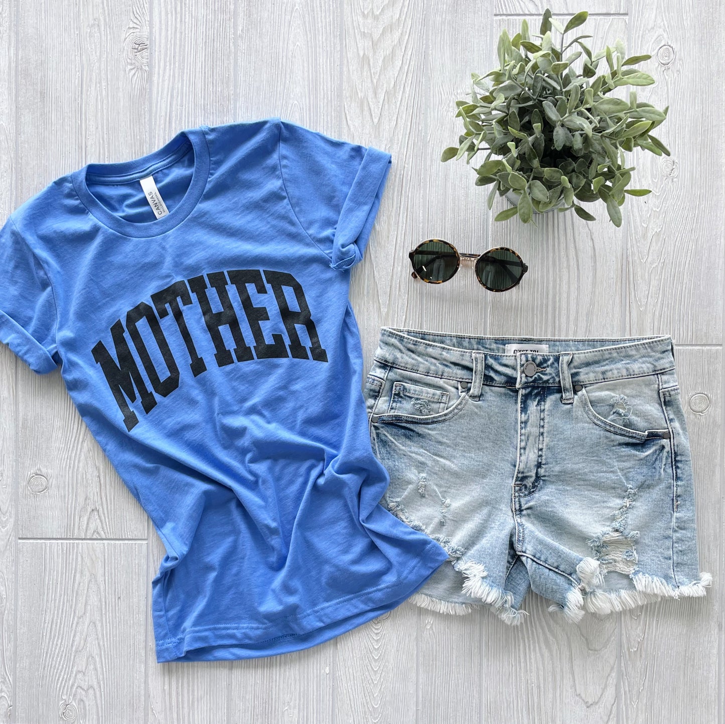 The MOTHER • Columbia Tee, a blue unisex Bella Canvas shirt with “MOTHER” in bold black letters, is styled with light denim cutoffs, round sunglasses, and a potted plant on a light wood floor for an easy everyday look.