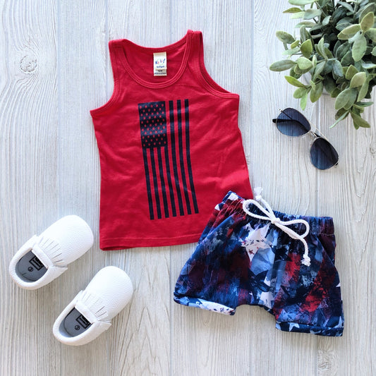 A flat lay of a toddler outfit featuring the Flag Tank • Kids, blue and red patterned shorts with a white drawstring, white slip-on shoes, black sunglasses, and a green plant on a light wooden floor.