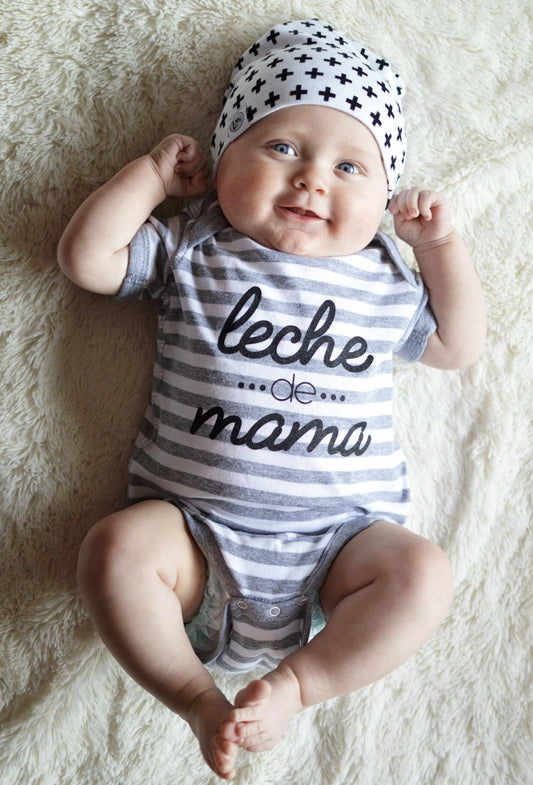 A smiling baby lies on a soft blanket, wearing the Leche de Mama Bodysuit—gender-neutral, hand-printed in Grand Rapids from 100% combed ring spun cotton—and a white hat with black plus signs.