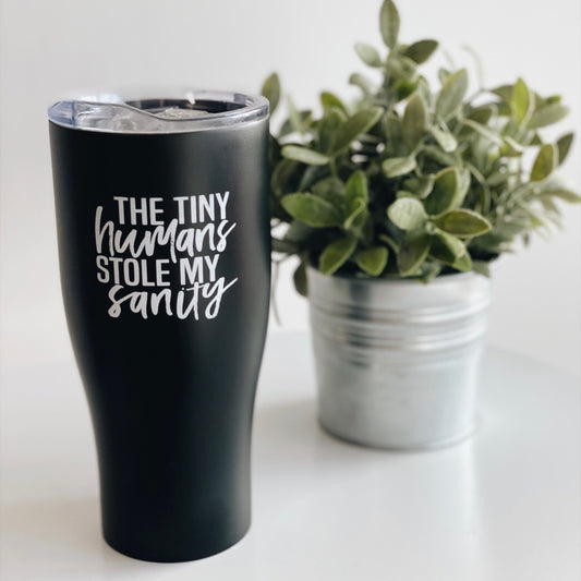 The Tiny Humans • Tumbler, a black stainless steel travel cup printed with The tiny humans stole my sanity, stands beside a potted green plant in a metal pot on a white surface.