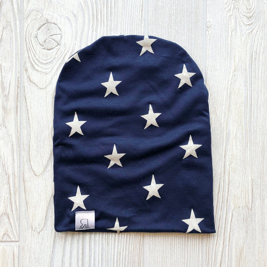 The Star Spangled Beanie is a navy blue handmade hat featuring a pattern of white stars and a small white tag with a black logo at the bottom edge, displayed flat on a light wooden surface.