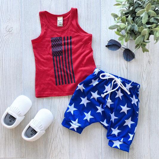 A super-soft Flag Tank • Kids with a blue American flag print, paired with blue star shorts, white slip-on shoes, black sunglasses, and a potted plant arranged on a light wooden floor.